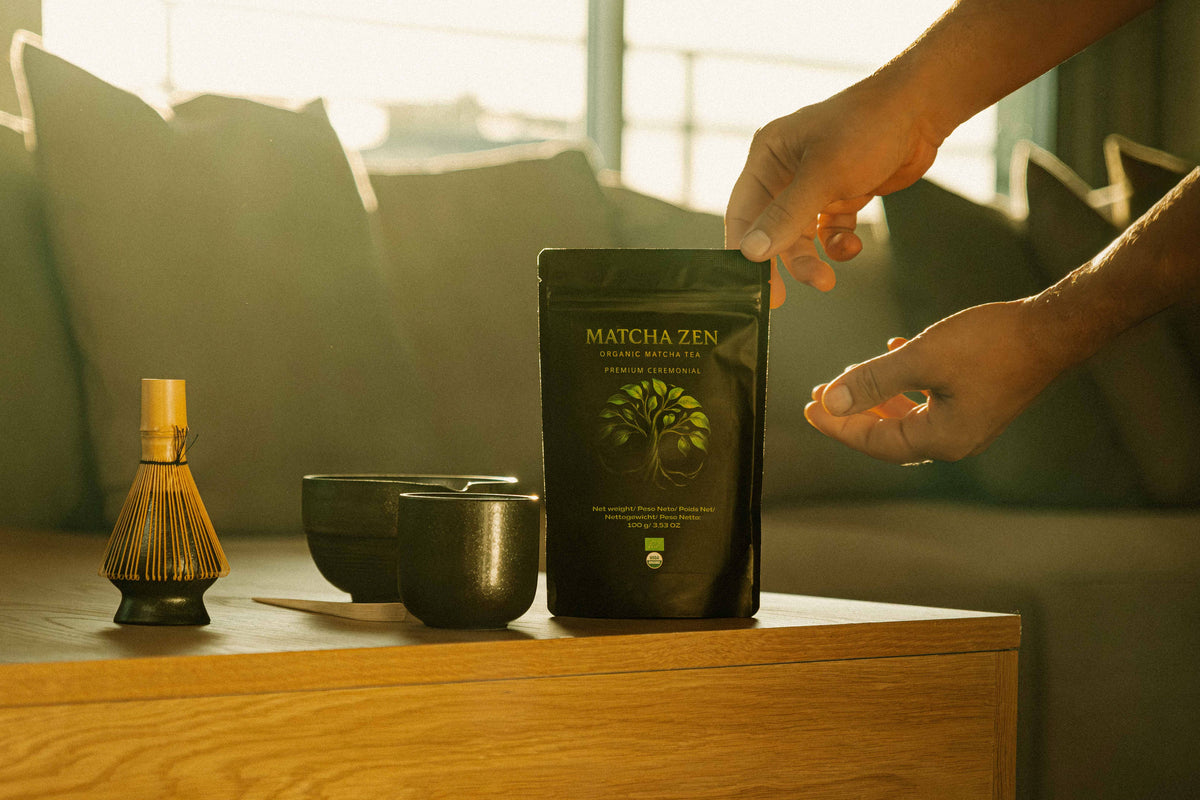 Manos tomando una bolsa de Matcha Zen junto a un chasen y un cuenco, con luz cálida de atardecer